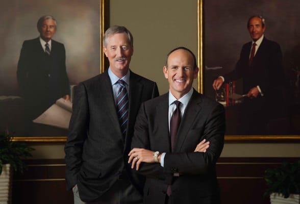 Steve Van Andel y Doug DeVos parados frente a retratos pintados de Jay Van Andel y Rich DeVos.